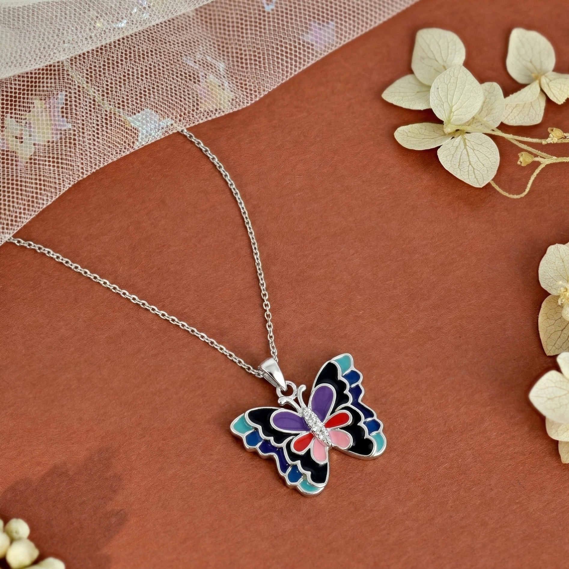 Handcrafted 925 sterling silver butterfly pendant necklace featuring intricate multi-color enamel wings in blue, purple, red, pink, and black with a central cubic zirconia stone body. Displayed on a textured brown background with dried white flowers and net tulle, this curated nature-inspired setup showcases romantic and stylish jewelry.