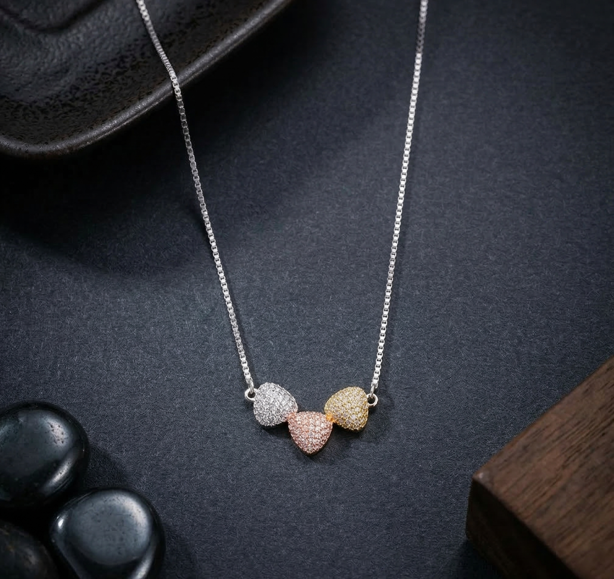 A 925 sterling silver necklace featuring a clustered pendant of three pavé-set heart motifs in silver, rose gold, and yellow gold finishes. The attached silver box chain rests on a dark textured slate background, elegantly styled alongside black polished stones, a geometric metal object, and dark wood accents. Modern minimalist Indian D2C jewelry, mixed metal styling, high-polish finish.