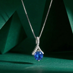 A delicate 925 sterling silver necklace suspended in mid-air, featuring a brilliant round deep blue center stone held by an elegant, sweeping ribbon-like pavé bail. Displayed against a modern, dark green geometric background with soft, focused lighting. Minimalist silver jewelry by CHAKORE, simulated sapphire pendant, everyday luxury, high-polish rhodium finish
