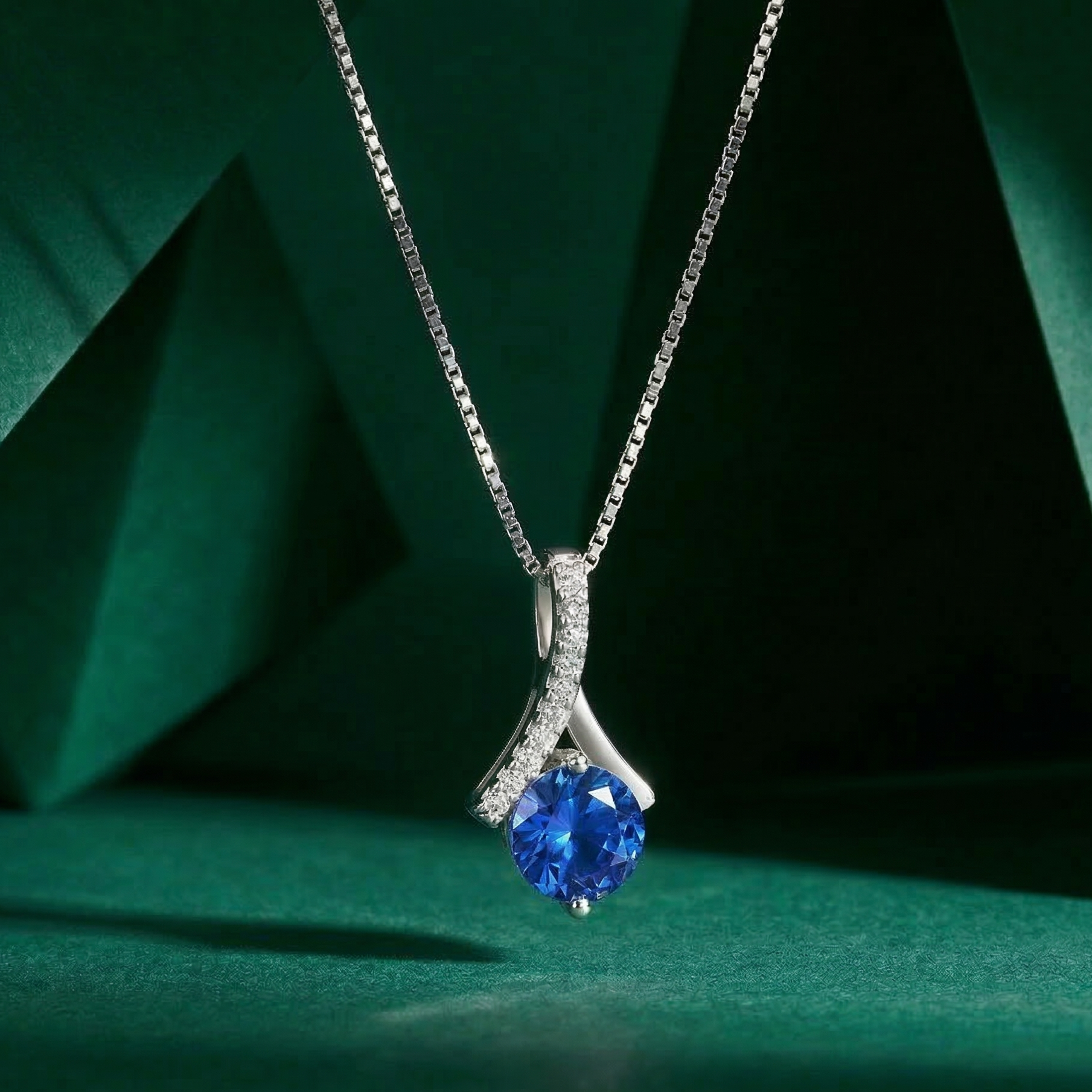 A delicate 925 sterling silver necklace suspended in mid-air, featuring a brilliant round deep blue center stone held by an elegant, sweeping ribbon-like pavé bail. Displayed against a modern, dark green geometric background with soft, focused lighting. Minimalist silver jewelry by CHAKORE, simulated sapphire pendant, everyday luxury, high-polish rhodium finish