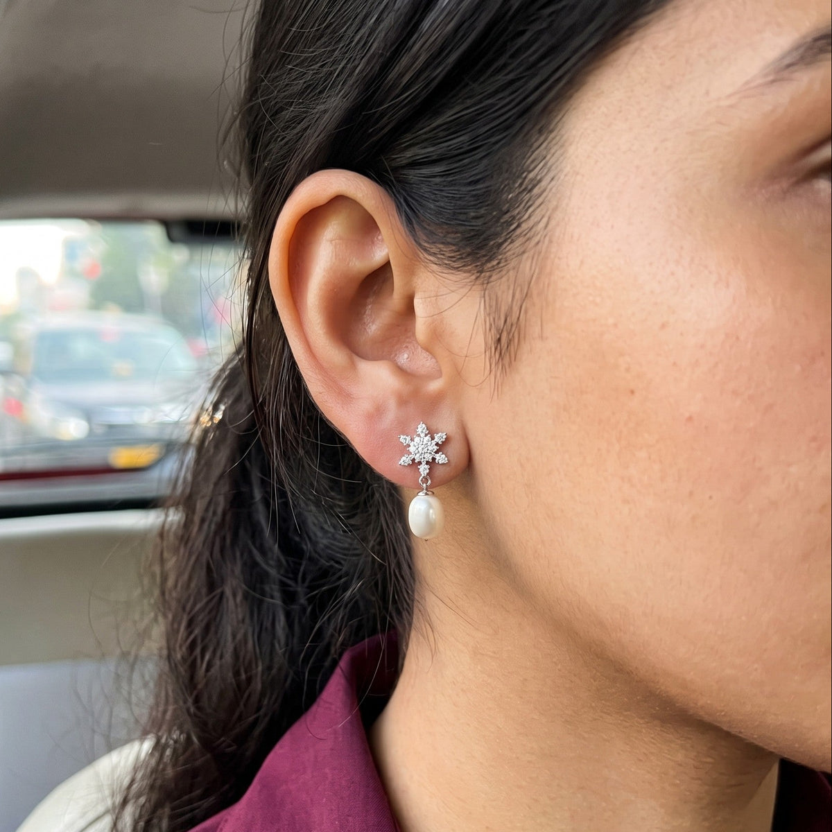 A close-up of a woman's ear wearing a 925 silver flower-shaped stud earring by Chakore with a white pearl drop, photographed inside a car.