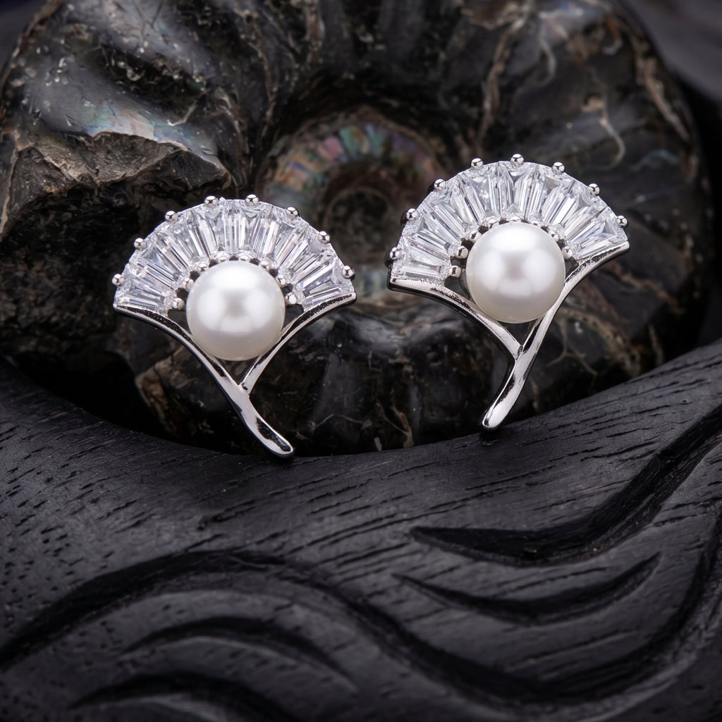 A pair of 925 pure silver fan-shaped stud earrings by Chakore with clear baguette crystals and a white pearl at the center, displayed on a dark carved wooden surface with a black shell behind them.