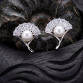 A pair of 925 pure silver fan-shaped stud earrings by Chakore with clear baguette crystals and a white pearl at the center, displayed on a dark carved wooden surface with a black shell behind them.