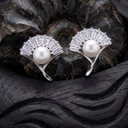A pair of 925 pure silver fan-shaped stud earrings by Chakore with clear baguette crystals and a white pearl at the center, displayed on a dark carved wooden surface with a black shell behind them.