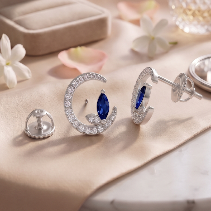 A 925 sterling silver crescent-shaped stud earrings by CHAKORE with marquise-cut blue gemstones and rows of clear crystals, displayed on a beige fabric surface with a ring box, perfume bottle, rose, flowers, and a hand mirror.The studs are anti-tarnish, rhodium polished and hypoallergenic.