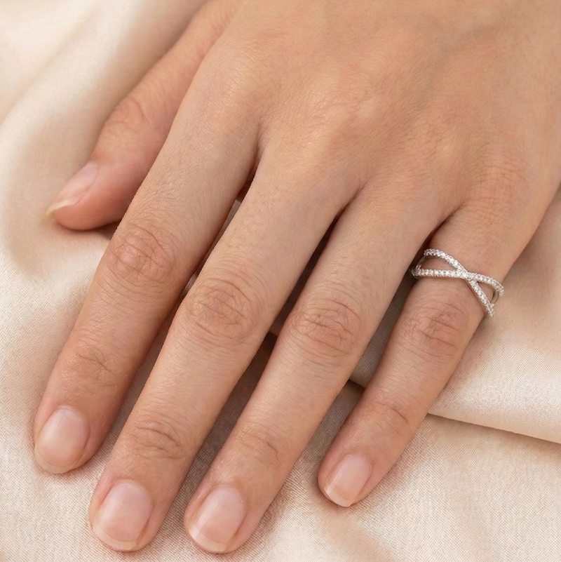 A close-up of a woman's hand wearing a delicate 925 sterling silver criss-cross ring by CHAKORE featuring pave cubic zirconia stones. The hand is resting elegantly on a smooth, soft peach-toned silk fabric backdrop. Minimalist silver jewelry, everyday wear stacking rings, high-polish finish.