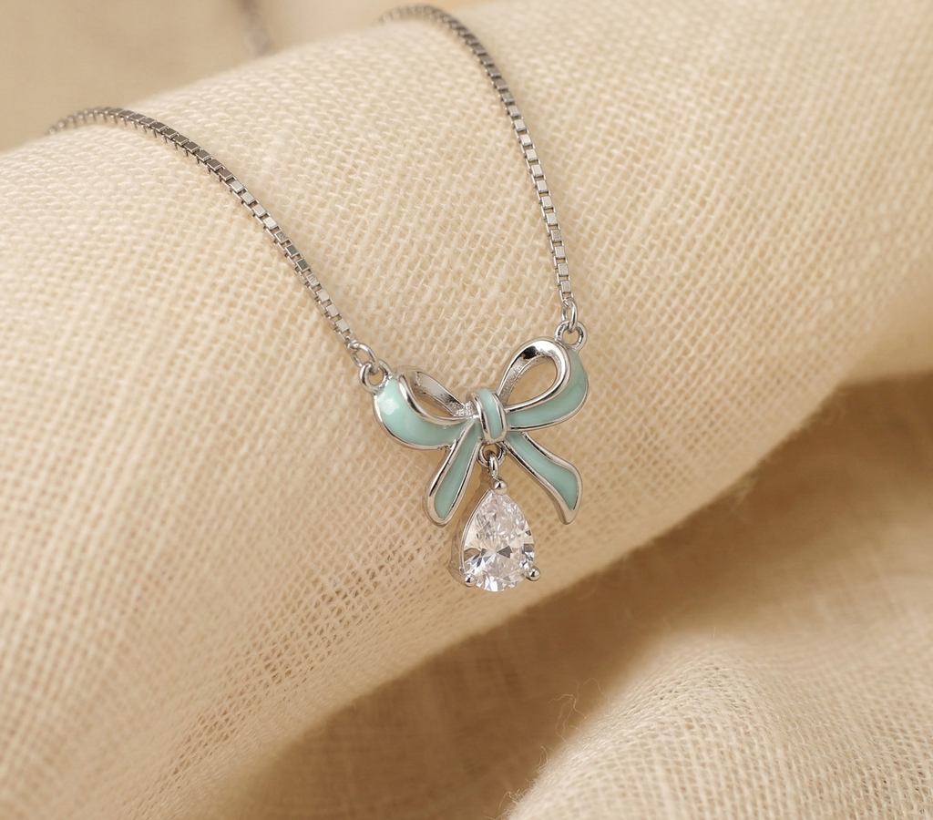 A delicate 925 sterling silver necklace featuring a mint green enamel bow pendant with a dangling pear-shaped cubic zirconia stone. The jewelry rests softly on a textured, light beige linen fabric. Modern coquette aesthetic silver jewelry, pastel enamel pendant, everyday luxury, high-polish rhodium finish.