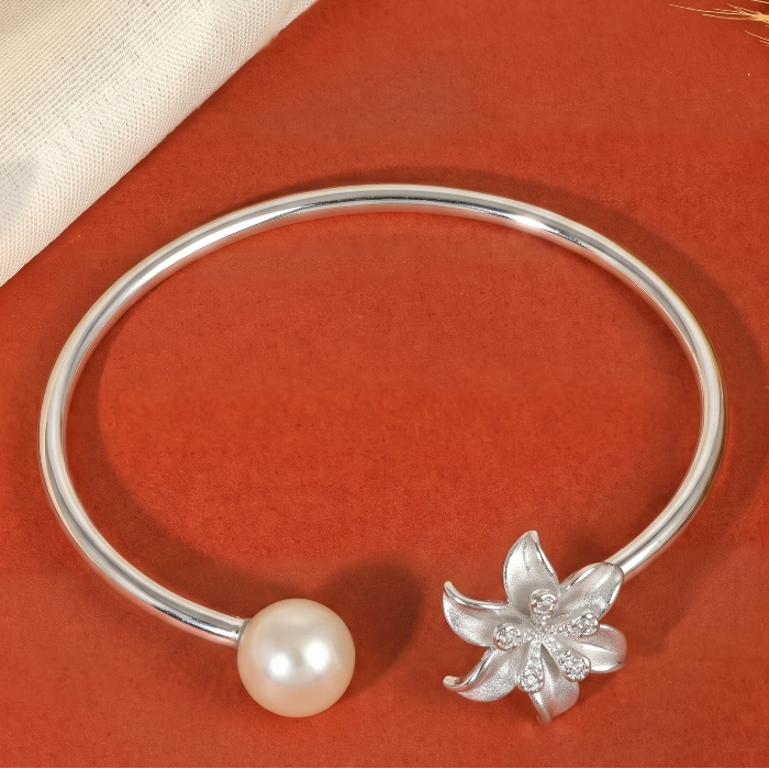 A silver-toned open bangle bracelet featuring a round white pearl and a silver flower charm set with small clear stones, displayed on a warm rust-orange background with cream fabric and dried grass accents.