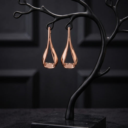 A pair of teardrop-shaped copper earrings hanging from a black branch-shaped jewelry display stand on a dark base, set against a dark paneled background.