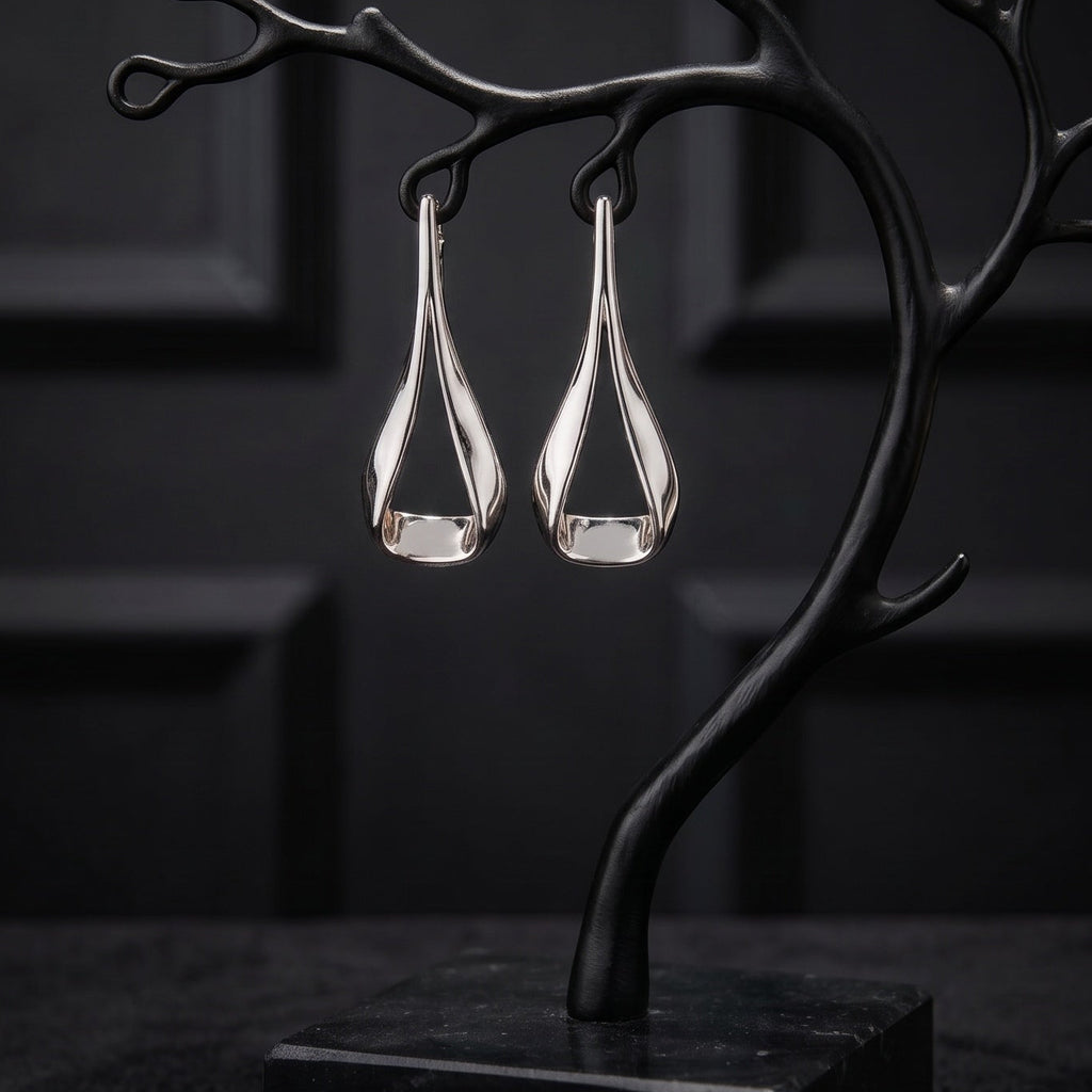 A pair of silver teardrop-shaped drop earrings displayed on a black branch-shaped jewelry stand with a square base, set against a dark paneled background.