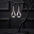 A pair of silver teardrop-shaped drop earrings displayed on a black branch-shaped jewelry stand with a square base, set against a dark paneled background.