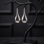 A pair of silver teardrop-shaped drop earrings displayed on a black branch-shaped jewelry stand with a square base, set against a dark paneled background.