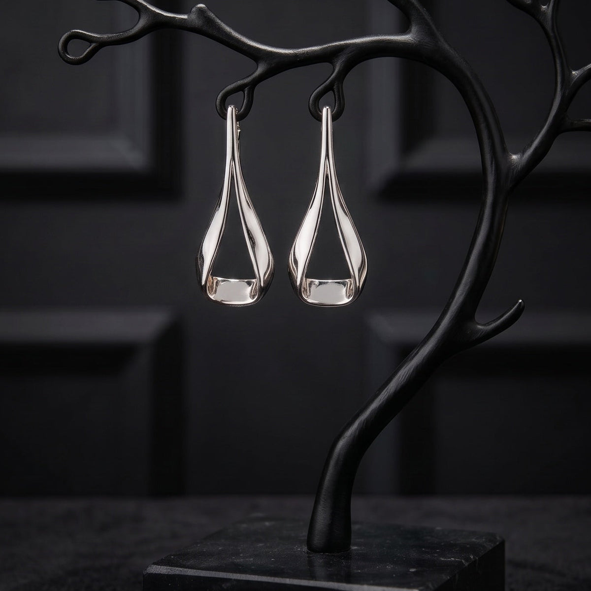 A pair of silver teardrop-shaped drop earrings displayed on a black branch-shaped jewelry stand with a square base, set against a dark paneled background.