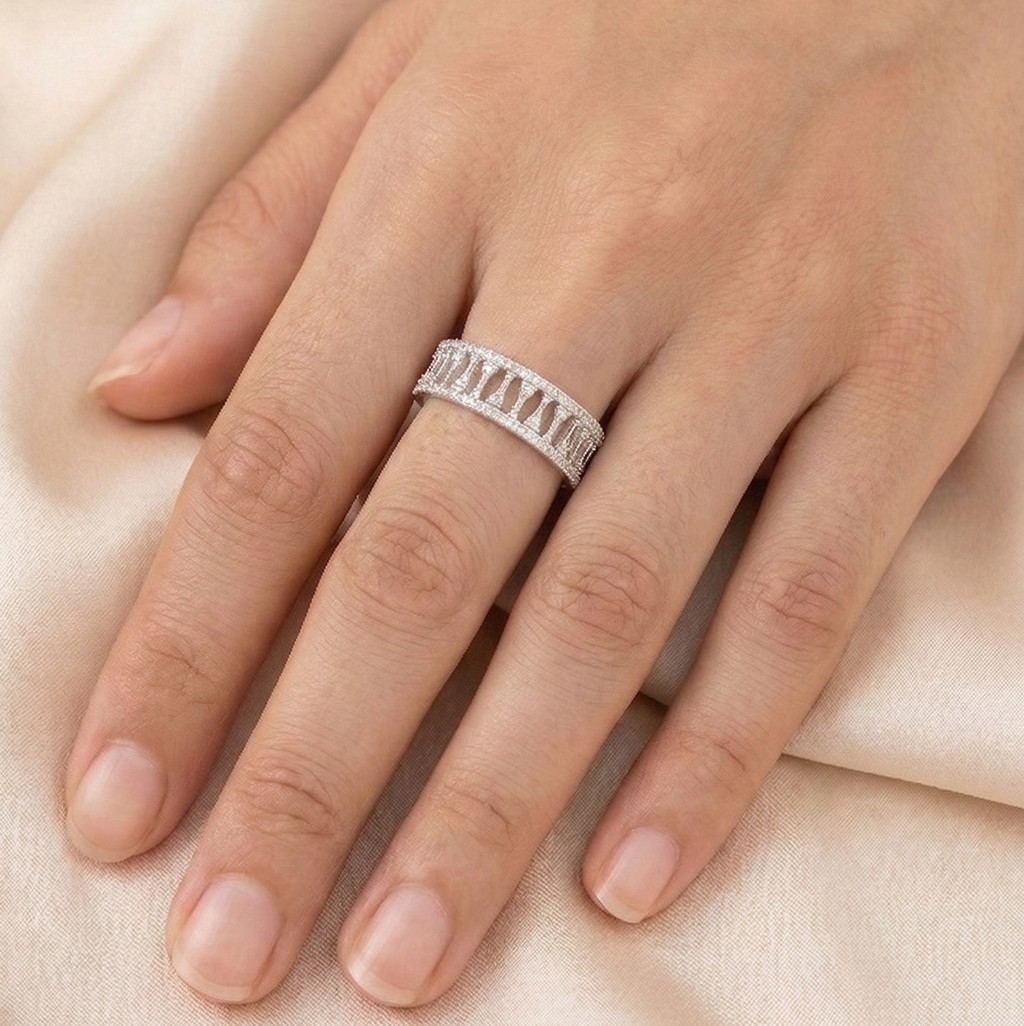 A close-up of a woman's hand resting elegantly on soft, draped peach silk fabric, modeling a wide 925 sterling silver statement ring. The ring features a repeating geometric cutout pattern bordered by delicate micro-pavé cubic zirconia stones. Modern minimalist silver jewellery, architectural wide band ring, high-polish everyday wear.