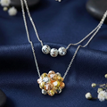 A trendy pre-layered 925 sterling silver necklace featuring diamond-cut textured beads. The shorter chain holds three silver beads, while the longer chain features a tri-color cluster sphere in silver, rose gold, and yellow gold. The jewelry is elegantly styled on draped midnight blue silk fabric, accented with smooth dark river stones and delicate white baby's breath flowers. Modern Indian D2C silver jewelry by CHAKORE, effortless layering, high-polish textured finish.