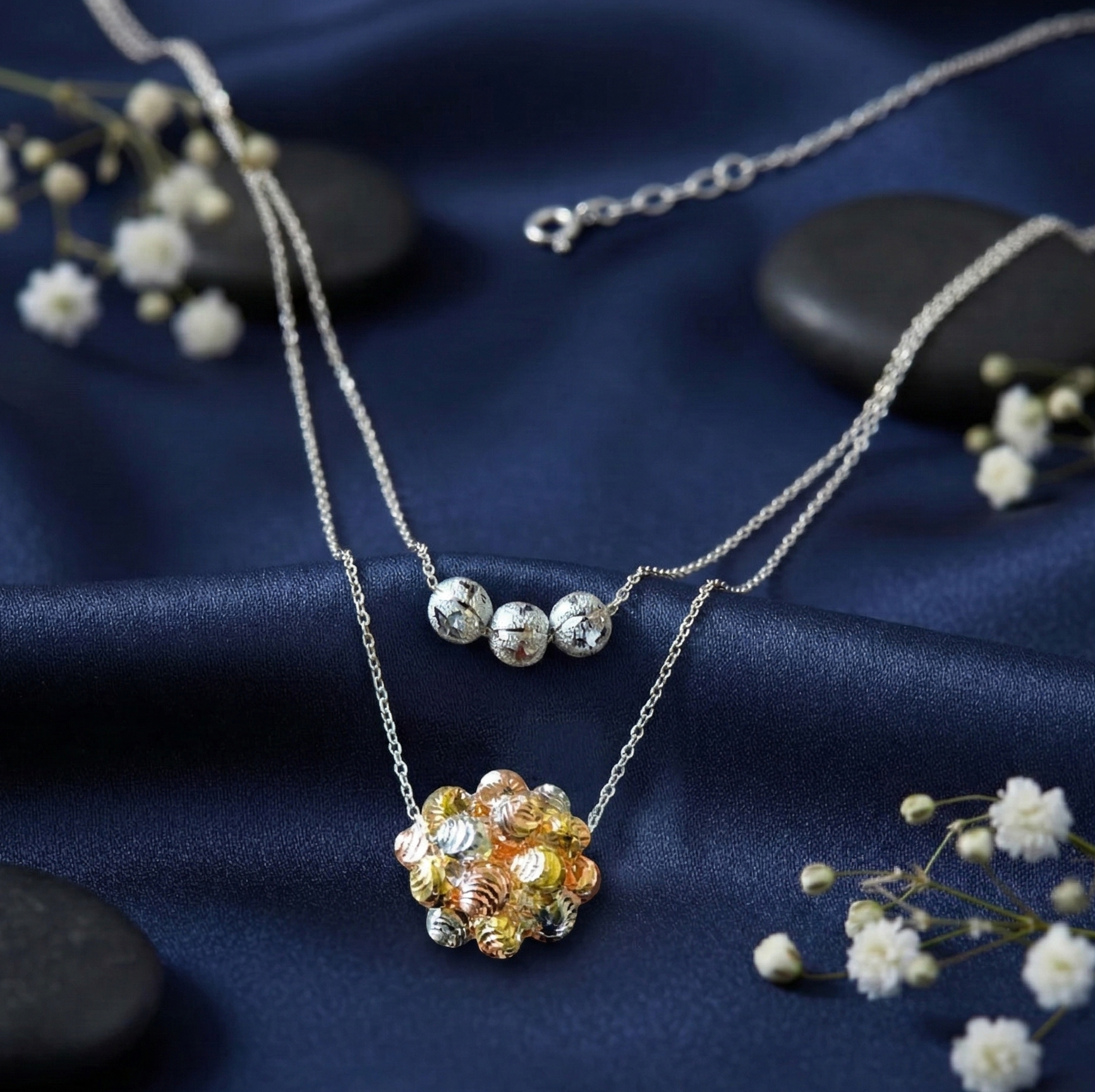 A trendy pre-layered 925 sterling silver necklace featuring diamond-cut textured beads. The shorter chain holds three silver beads, while the longer chain features a tri-color cluster sphere in silver, rose gold, and yellow gold. The jewelry is elegantly styled on draped midnight blue silk fabric, accented with smooth dark river stones and delicate white baby's breath flowers. Modern Indian D2C silver jewelry by CHAKORE, effortless layering, high-polish textured finish.