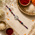 A traditional Indian 925 silver Rakhi by CHAKORE with a silver-toned circular charm engraved with Devanagari script, flanked by red and blue beads and silver-toned spacer beads. The rakhi is displayed diagonally on a beige embroidered cloth surrounded by festive props including bowls of red powder and white rice.