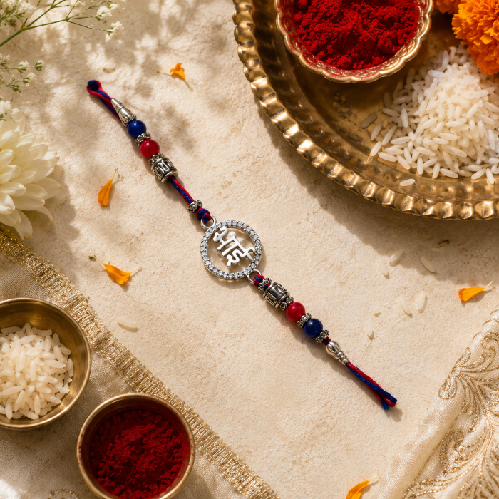 A traditional Indian 925 silver Rakhi by CHAKORE with a silver-toned circular charm engraved with Devanagari script, flanked by red and blue beads and silver-toned spacer beads. The rakhi is displayed diagonally on a beige embroidered cloth surrounded by festive props including bowls of red powder and white rice.