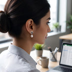 A woman in a light blue collared shirt sits at a wooden desk, looking at a laptop computer in a bright, modern office setting wearing a 925 silver stud earrings with real pearl drop by Chakore. 