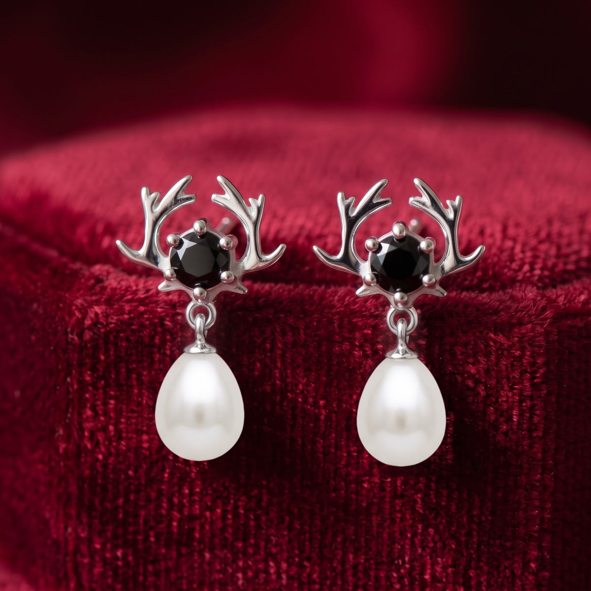 A pair of 925 silver stud earrings by Chakore with antler-shaped tops, black round stones, and dangling white teardrop pearls, displayed on a deep red velvet surface.