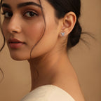 A close-up portrait of a woman wearing a small 925 sterling silver butterfly-shaped stud earring by CHAKORE with clear stones, set against a warm beige background. The studs are anti-tarnish, rhodium polished and hypoallergenic pure 925 silver.