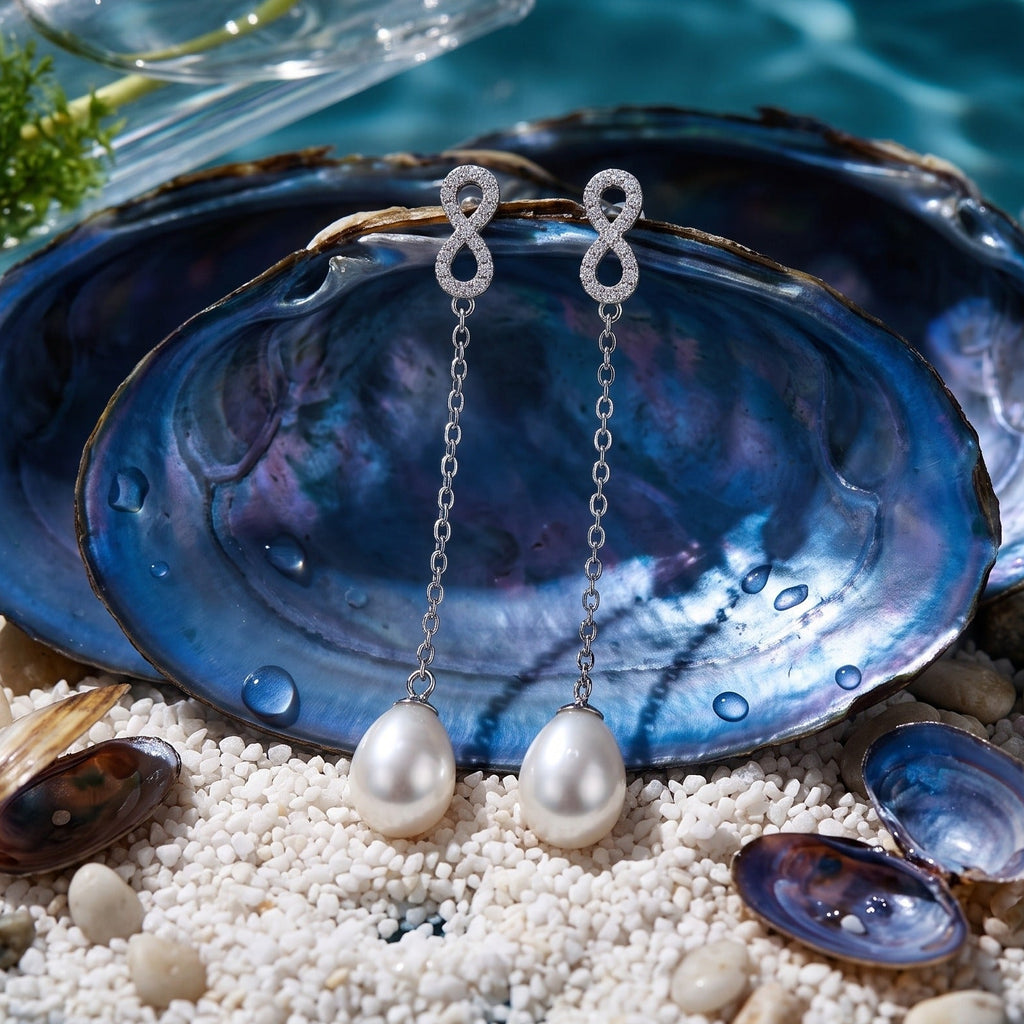 A pair of 925 silver drop earrings by Chakore with oval white freshwater pearls and sparkling crystal-like top details, displayed on blue abalone shells over white pebbles with turquoise water in the background.