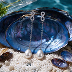 A pair of 925 silver drop earrings by Chakore with oval white freshwater pearls and sparkling crystal-like top details, displayed on blue abalone shells over white pebbles with turquoise water in the background.