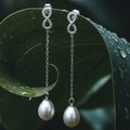 CHAKORE 925 sterling silver infinity earrings with genuine freshwater pearl drops, hanging from a curled green leaf covered in raindrops.
