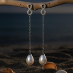 CHAKORE anti-tarnish 925 sterling silver infinity freshwater pearl earrings hanging from driftwood above dark sand and shells, with blue water behind.