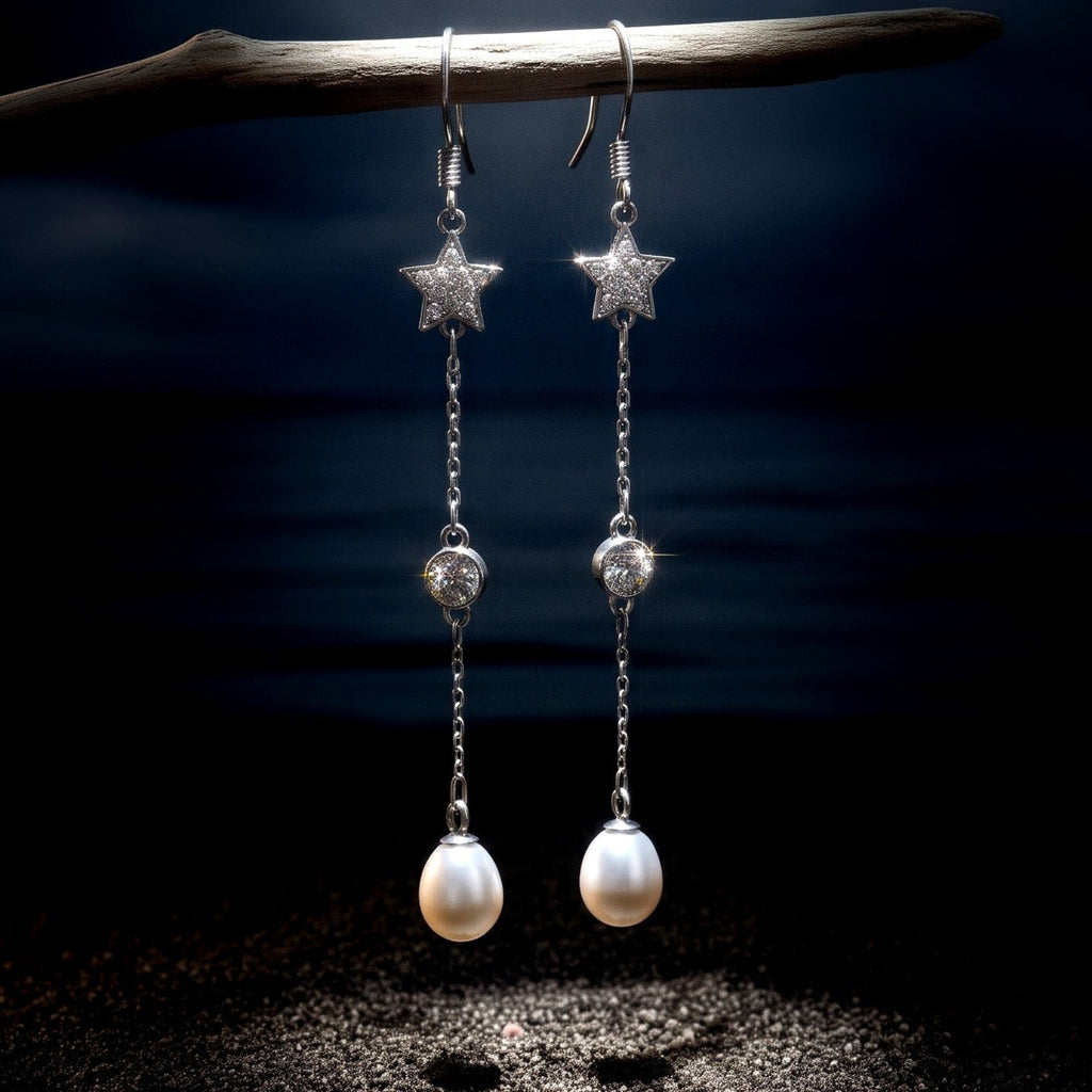 A pair of 925 silver dangle earrings by Chakore with star-shaped tops, clear faceted beads, and white freshwater pearl drops, hanging from a dark branch over a beach-like setting with sand, shells, and a dark water background. 