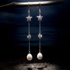 A pair of 925 silver dangle earrings by Chakore with star-shaped tops, clear faceted beads, and white freshwater pearl drops, hanging from a dark branch over a beach-like setting with sand, shells, and a dark water background. 