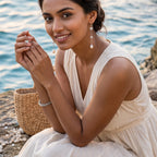 A woman in a cream-colored sleeveless dress sits on a rocky shore with the ocean in the background, wearing a 925 silver long freshwater pearl drop earrings by Chakore silver jewellery and a bracelet. 