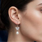 A close-up of a woman's ear wearing a 925 silver dangle earring by Chakore with a small floral cluster of clear stones and a dangling white pearl, against a dark blue background.