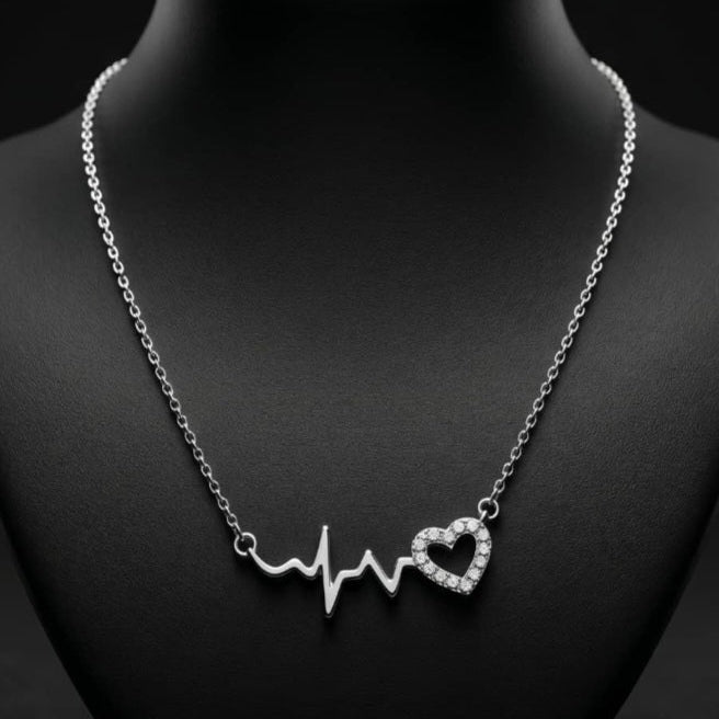Front view of a CHAKORE rhodium-plated 925 silver necklace displayed on a black jewellery bust against a dark background. The necklace features a central pendant with a smooth polished heartbeat line that connects to an open heart shape encrusted with clear pave crystals, suspended on a delicate silver cable chain.