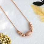 A 925 sterling silver necklace with a rose gold finish, featuring a free-sliding cluster of beads strung directly onto a delicate chain. The center is a frosted oval bead flanked by smooth and diamond-cut spherical beads. The jewelry rests gracefully on a textured white fabric background, accented by a dried yellow fern, dark green leaves, and a small cluster of dried white flowers. Minimalist Indian D2C jewelry, rose gold slider necklace, everyday modern styling.