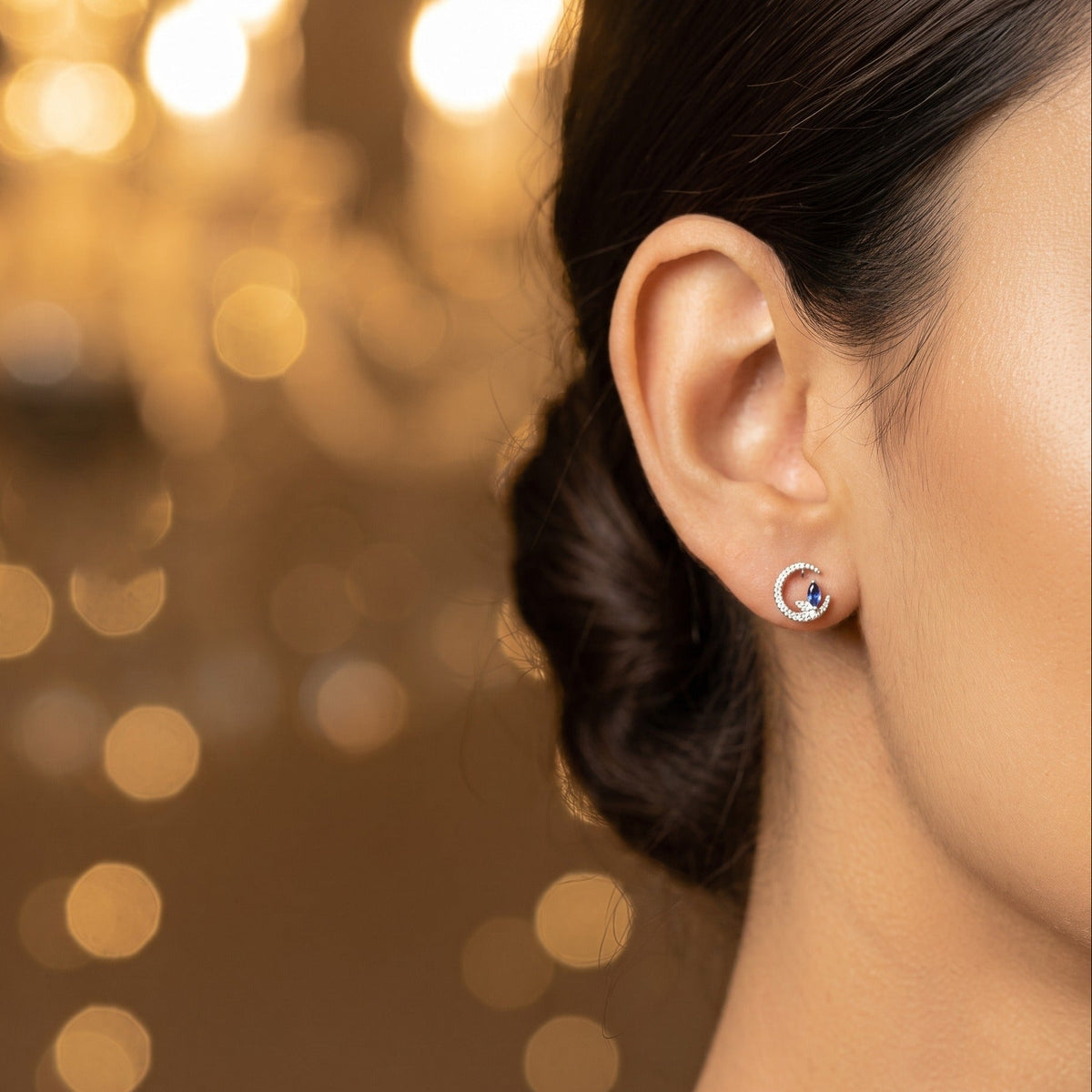 A close-up side profile of a woman wearing a small silver hoop earring by Chakore with a round blue gemstone, set against a warm, softly blurred background with golden bokeh lights.