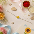 A delicate 925 silver chain bracelet rakhi by CHAKORE with a yellow daisy charm featuring a pink center, styled in a flat-lay arrangement with wooden stars, a candle, rice, red powder, and ribbons on a light beige background.